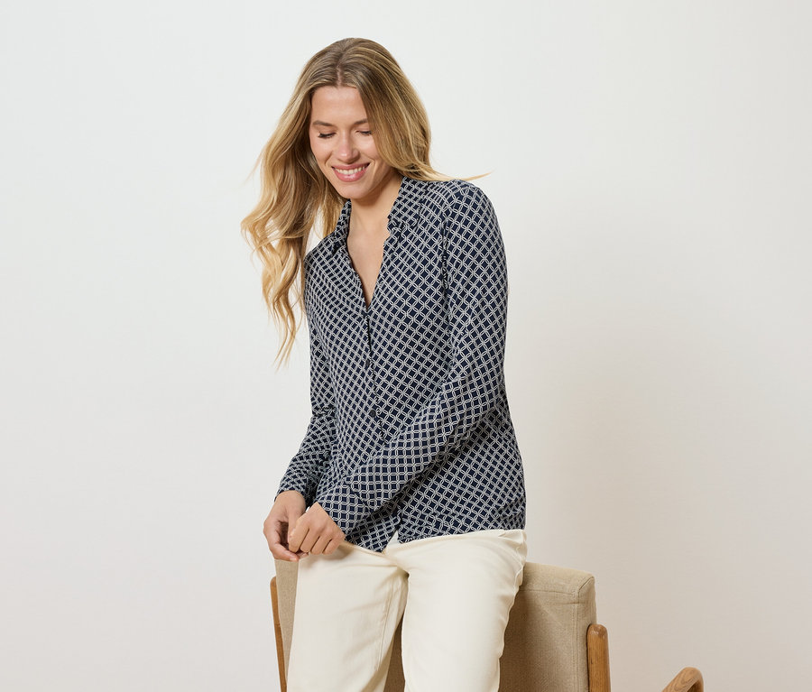 Une femme aux longs cheveux blonds, portant un chemisier à motifs et un pantalon clair, est assise sur une chaise et sourit.