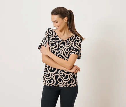 Une femme aux bras croisés porte un chemisier à motifs noirs et beiges et un pantalon noir.
