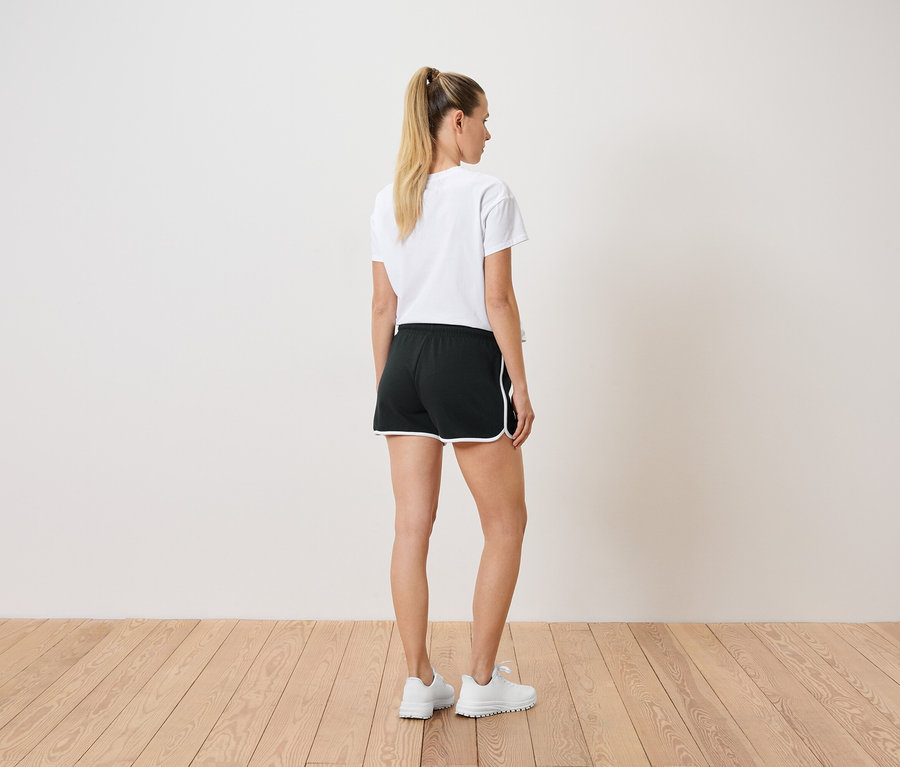 Une femme avec une queue de cheval porte un t-shirt blanc, un short noir avec des bordures blanches et des baskets blanches, vue de dos.