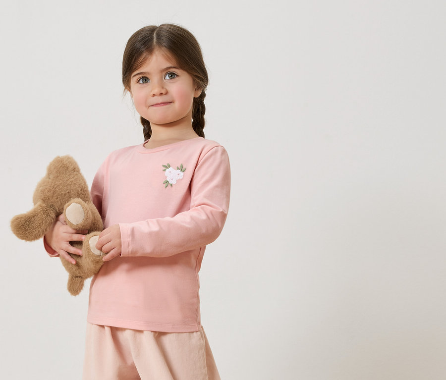 Fillette avec des tresses tenant un ours en peluche, portant un t-shirt rose à manches longues avec une application florale et un short rose.