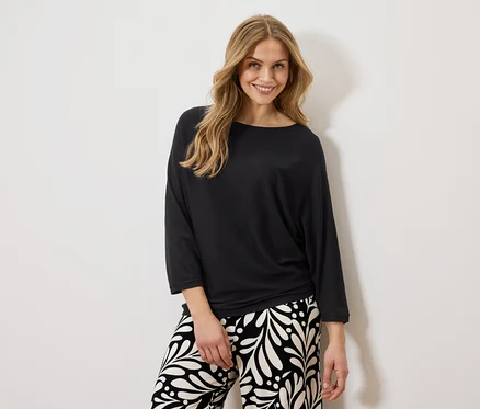 Femme souriante portant un haut noir et un pantalon à motifs devant un mur blanc.