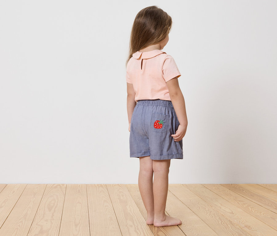 Fillette vue de dos, portant une chemise rose et un short bleu avec une application fraise.