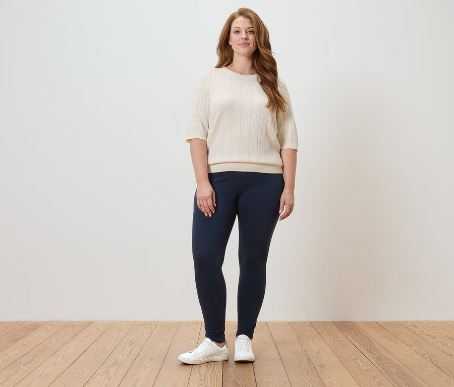 Une femme se tient dans une pièce lumineuse, portant un pull crème, un legging bleu marine et des baskets blanches.