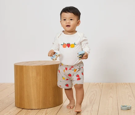 Un jeune enfant se tient pieds nus sur un plancher en bois et tient deux petites voitures jouets dans ses mains. Il porte un haut à manches longues avec des motifs de fruits et un short court avec un motif de fraises.