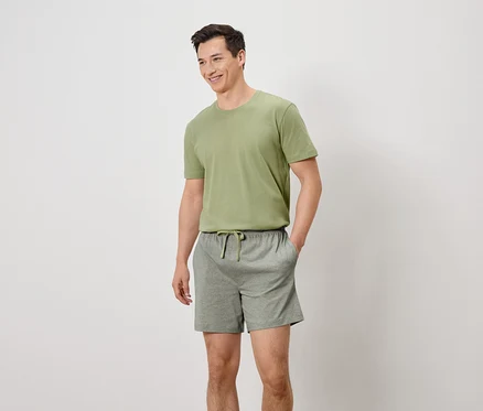 Homme souriant portant un t-shirt vert olive et un short gris avec cordon de serrage, les mains dans les poches.