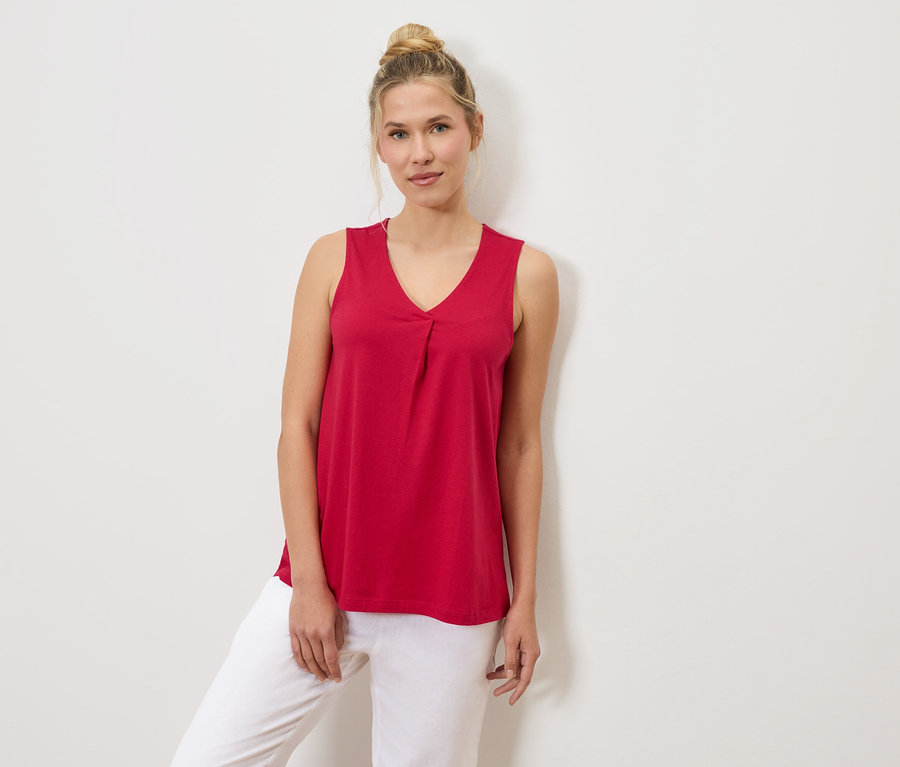 Une femme portant un chignon est vêtue d'un débardeur rouge et d'un pantalon blanc, appuyée contre un mur blanc.