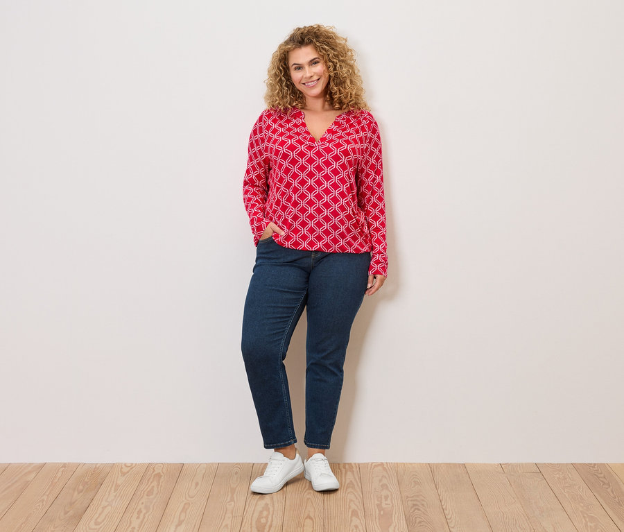 Femme aux cheveux blonds bouclés, chemisier rouge à motifs blancs, jean et baskets blanches, debout devant un mur.