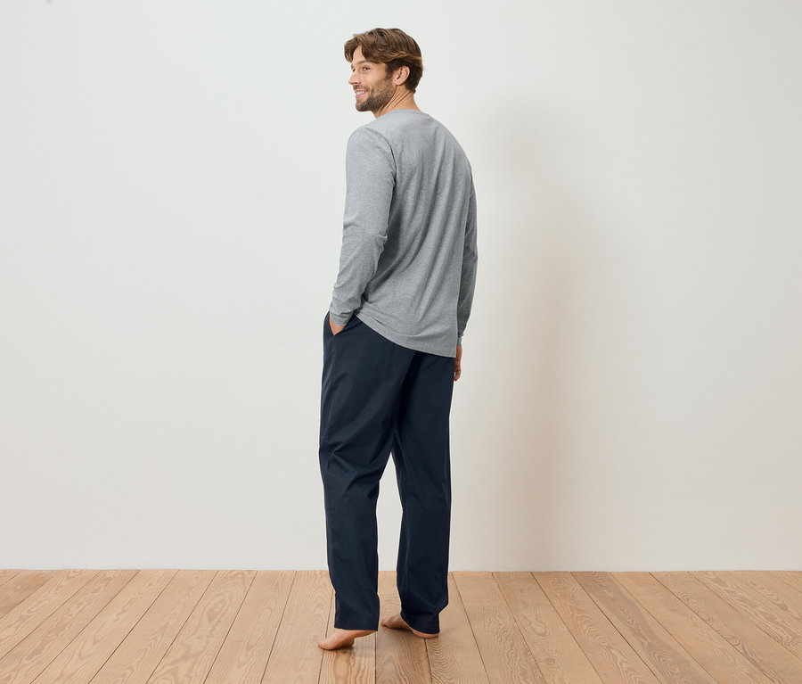 Un homme barbu porte un t-shirt gris à manches longues et un pantalon bleu marine, les mains dans les poches, regardant par-dessus son épaule.