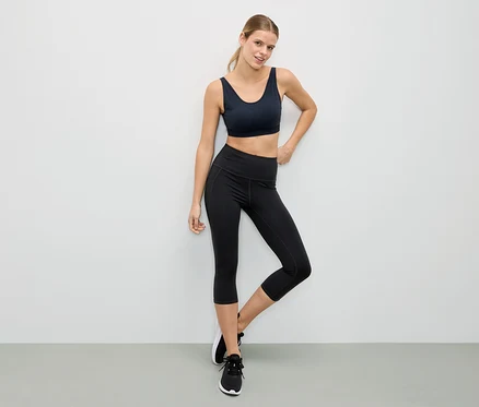 Femme posant en soutien-gorge de sport noir et leggings noirs devant un mur blanc.