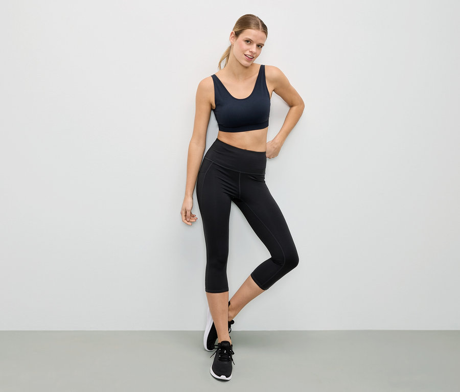 Femme posant en soutien-gorge de sport noir et leggings noirs devant un mur blanc.