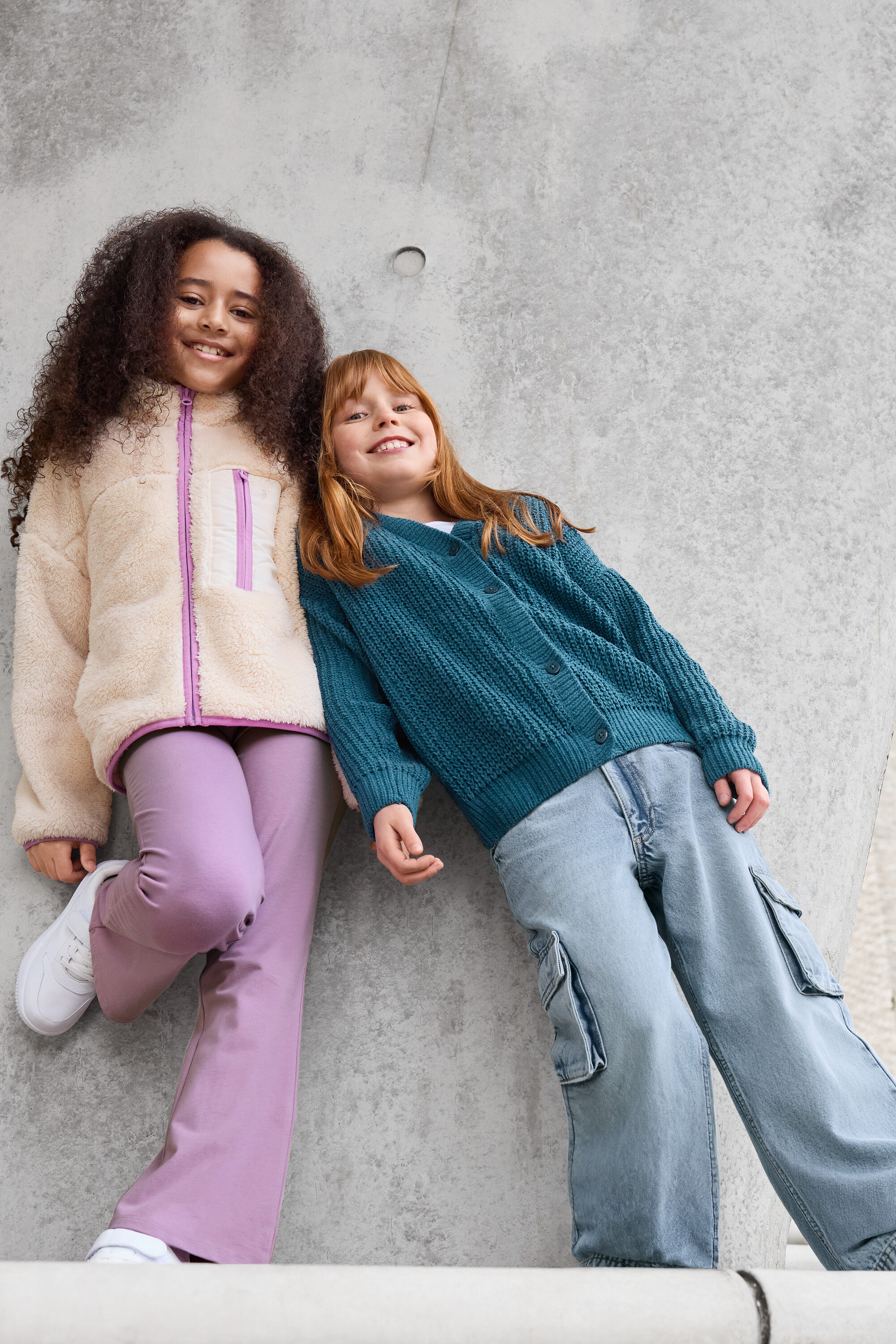 Deux filles sont appuyées contre un mur en béton. La fille de gauche porte une veste en teddy, un pantalon violet et des baskets blanches. La fille de droite porte un cardigan vert et un pantalon bleu.
