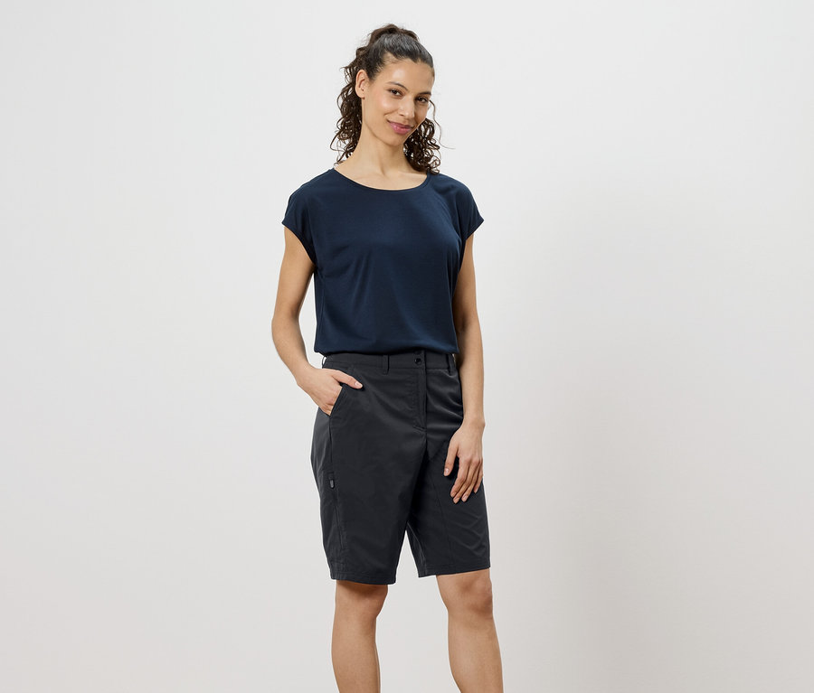 Une femme aux cheveux bruns bouclés porte un t-shirt bleu marine et un short gris, la main dans la poche.