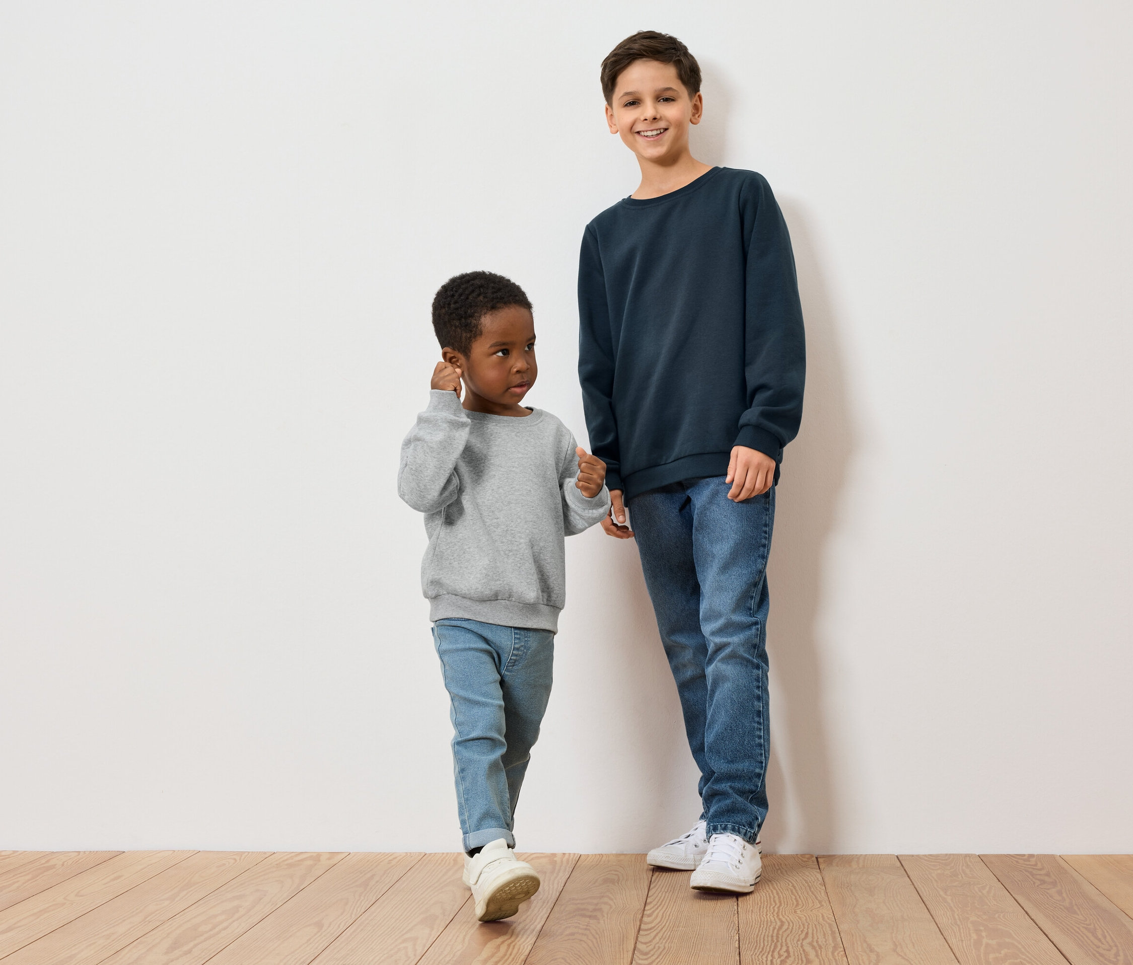 Deux garçons, un à la peau foncée en pull gris, l'autre à la peau claire en pull bleu, se tiennent sur un plancher en bois devant un mur blanc.
