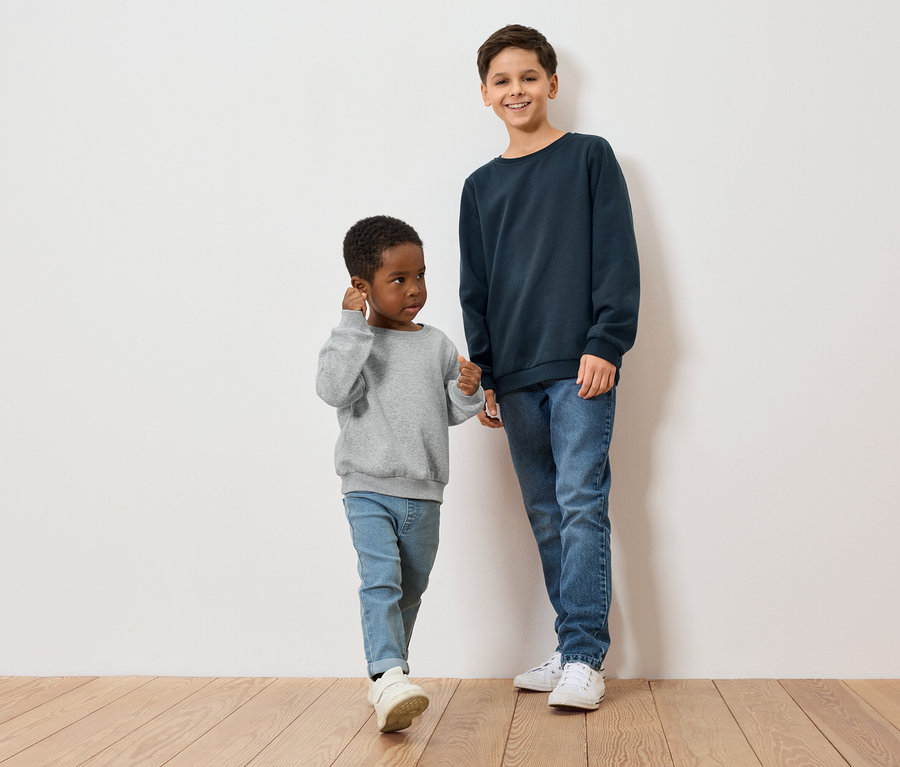 Deux garçons, un à la peau foncée en pull gris, l'autre à la peau claire en pull bleu, se tiennent sur un plancher en bois devant un mur blanc.