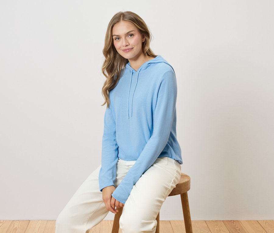 Une femme aux longs cheveux blonds est assise sur une chaise en bois, portant un sweat à capuche bleu clair et un pantalon blanc.
