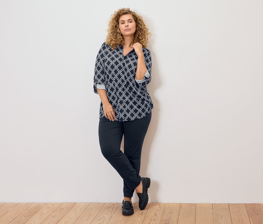 Femme aux cheveux blonds bouclés portant un chemisier à motifs bleu et blanc, un pantalon foncé et des chaussures noires, appuyée contre un mur.