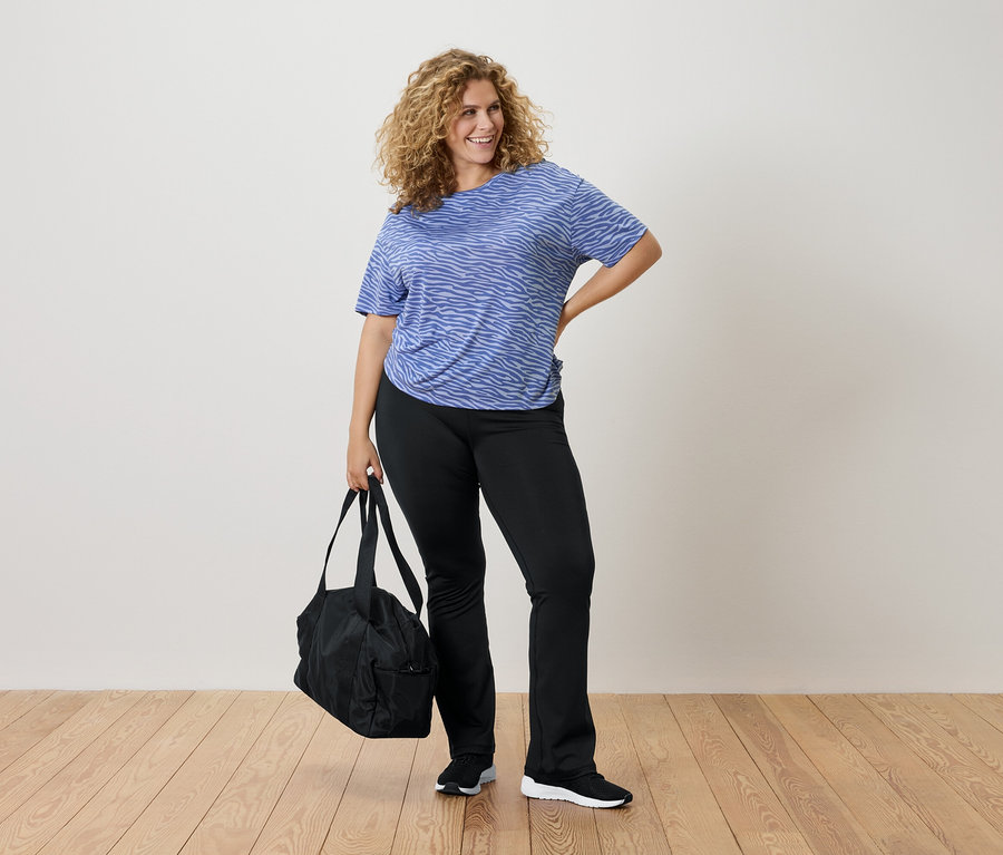 Femme aux cheveux bouclés portant un t-shirt zébré bleu, un pantalon noir et tenant un sac de sport noir.