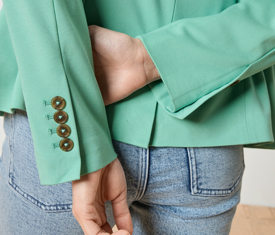 Gros plan d'une personne vue de dos portant une veste vert clair avec quatre boutons sur la manche et un jean.