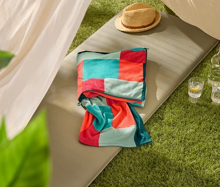 Une serviette de plage multicolore est posée sur un tapis de sol vert olive, à côté d'un chapeau de paille et de deux verres d'eau avec du citron.