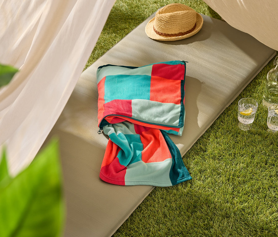 Une serviette de plage multicolore est posée sur un tapis de sol vert olive, à côté d'un chapeau de paille et de deux verres d'eau avec du citron.