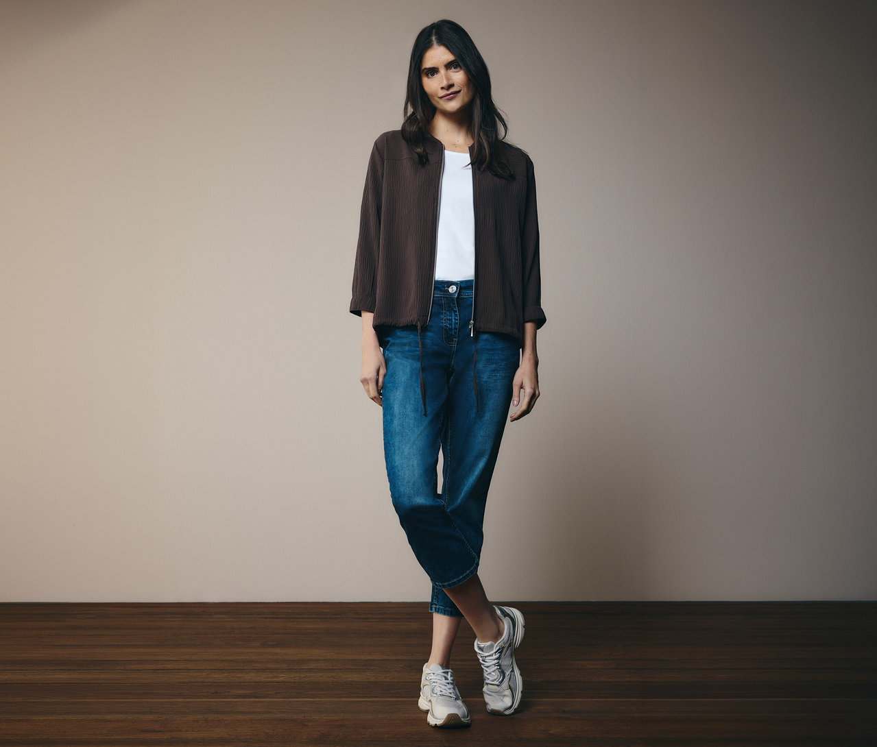 Femme portant un t-shirt blanc, un jean et un blouson CECIL marron. Elle se tient devant un mur.