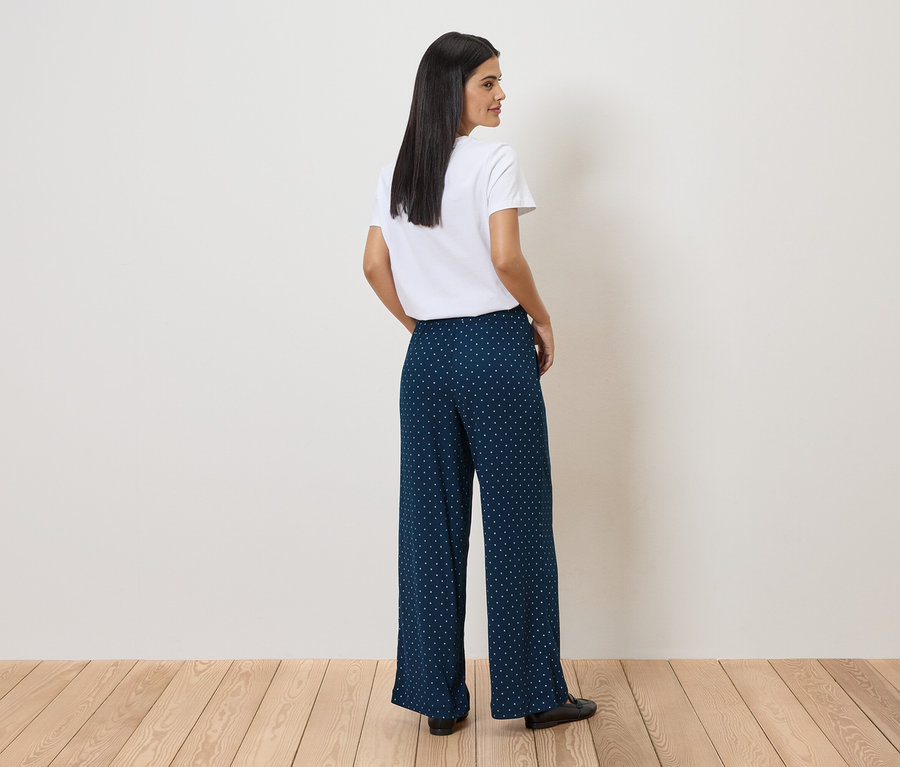 Une femme portant un t-shirt blanc et un pantalon bleu marine à pois blancs se tient sur un plancher en bois et regarde par-dessus son épaule.