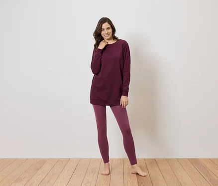 Une femme se tient pieds nus sur un plancher en bois devant un mur blanc et porte un long haut bordeaux et des leggings.