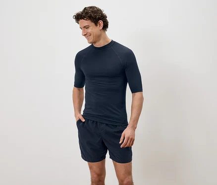 Un homme aux cheveux bouclés portant une chemise à manches courtes bleu marine et un short devant un fond uni.