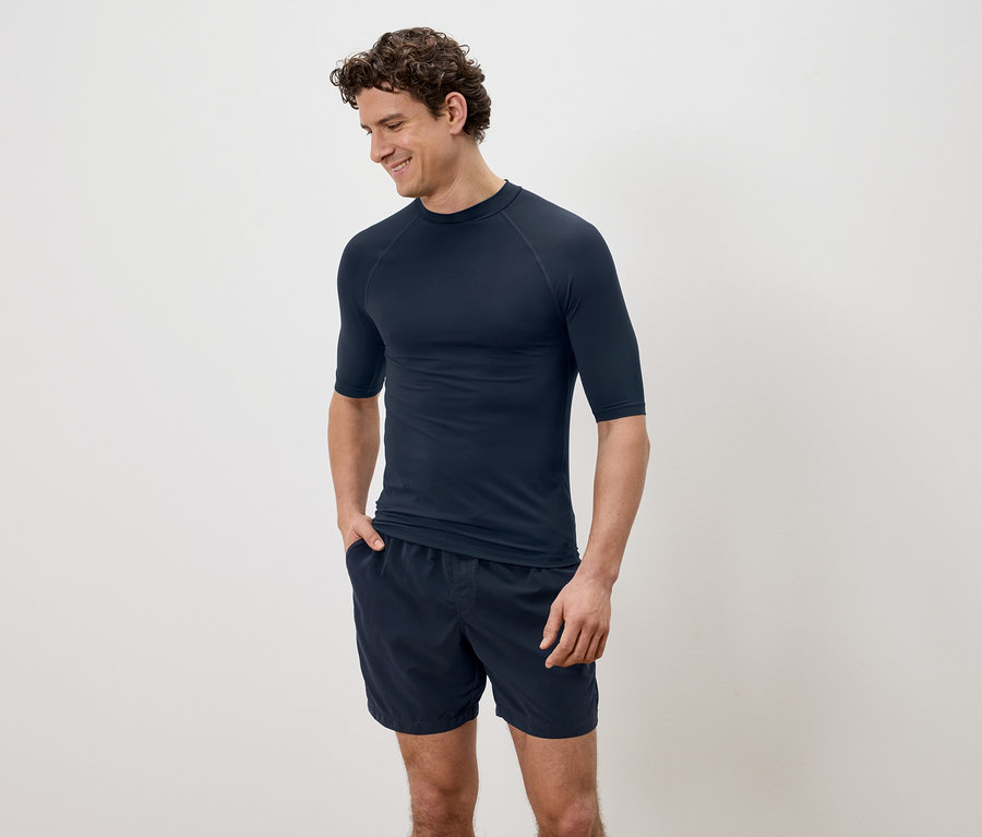 Un homme aux cheveux bouclés portant une chemise à manches courtes bleu marine et un short devant un fond uni.