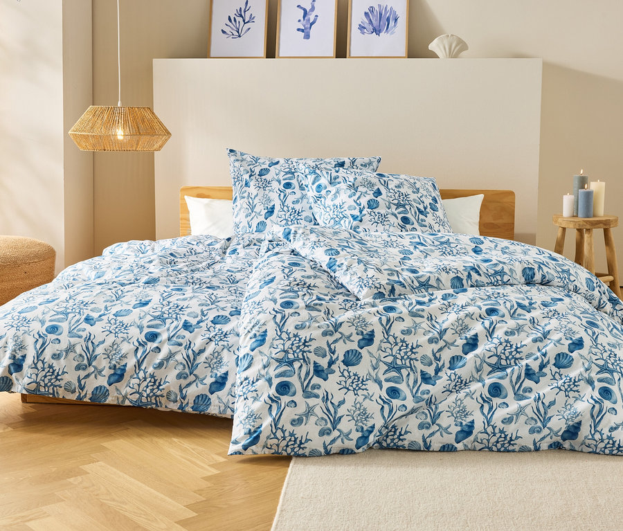 Un lit avec des draps à motifs bleus et blancs se trouve dans une chambre. Au-dessus du lit sont accrochés trois tableaux encadrés et une lampe.