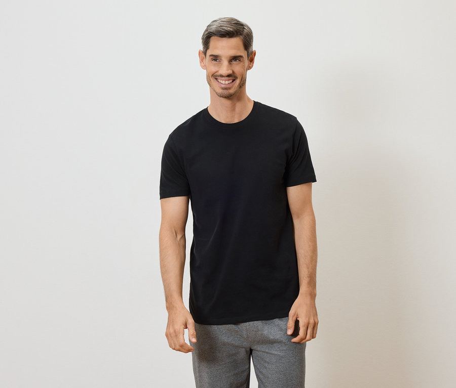 Un homme aux cheveux gris souriant porte un t-shirt noir et un pantalon gris sur fond blanc.