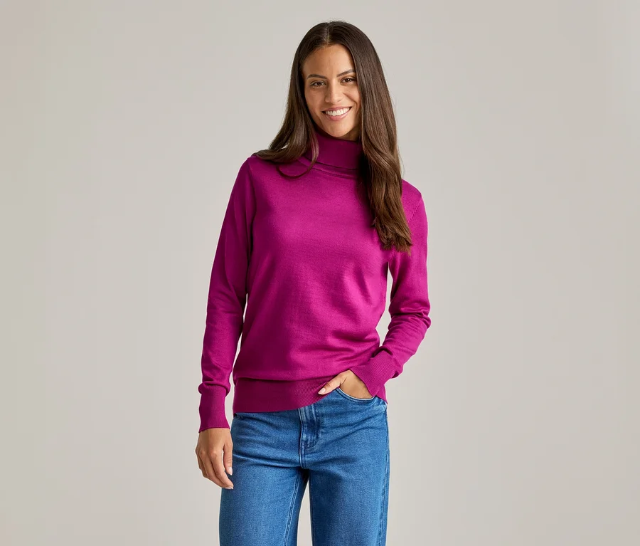 Une femme souriante portant un pull à col roulé rose et un jean bleu, une main dans la poche.