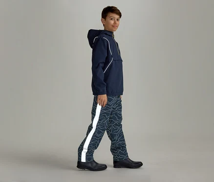 Un garçon portant un imperméable bleu marine à capuche et un pantalon de pluie à motifs avec une bande réfléchissante marche de côté vers la droite.