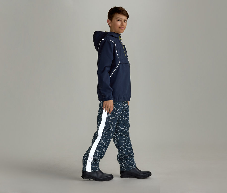 Un garçon portant un imperméable bleu marine à capuche et un pantalon de pluie à motifs avec une bande réfléchissante marche de côté vers la droite.
