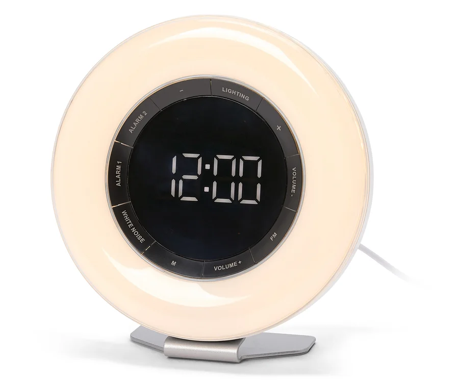Un réveil lumineux blanc et circulaire avec un anneau extérieur illuminé et un écran central sombre affichant "12:00". Des boutons de commande tels que "ALARM 1", "ALARM 2", "LIGHTING", "WHITE NOISE", "FM" et "VOLUME" sont visibles autour de l'écran. Le réveil repose sur une base argentée.