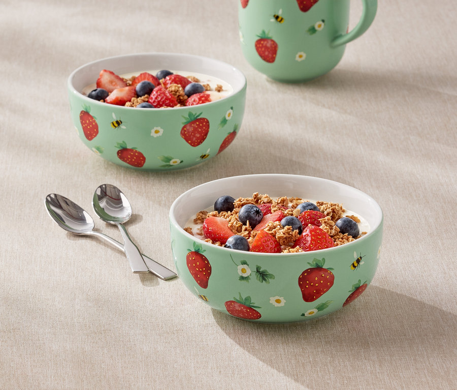 Deux bols remplis de yaourt, de fraises et de myrtilles avec un motif de fraises et un pichet sont posés sur une table.