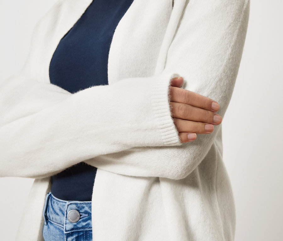Une femme aux bras croisés porte un long cardigan blanc sur un haut bleu marine et un jean bleu.