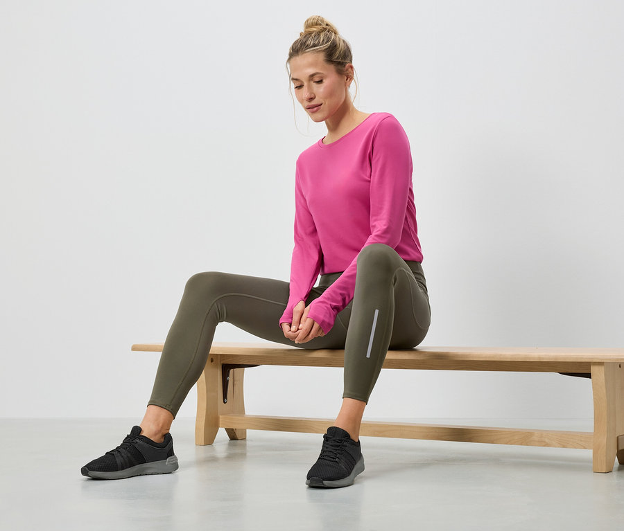 Femme assise sur un banc en bois, portant un haut rose à manches longues, un legging vert et des baskets noires.