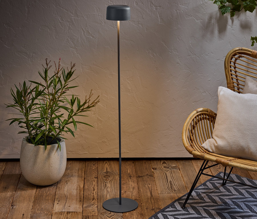 Un lampadaire se trouve à côté d'une plante en pot et d'une chaise en osier avec coussin sur un tapis à motifs.