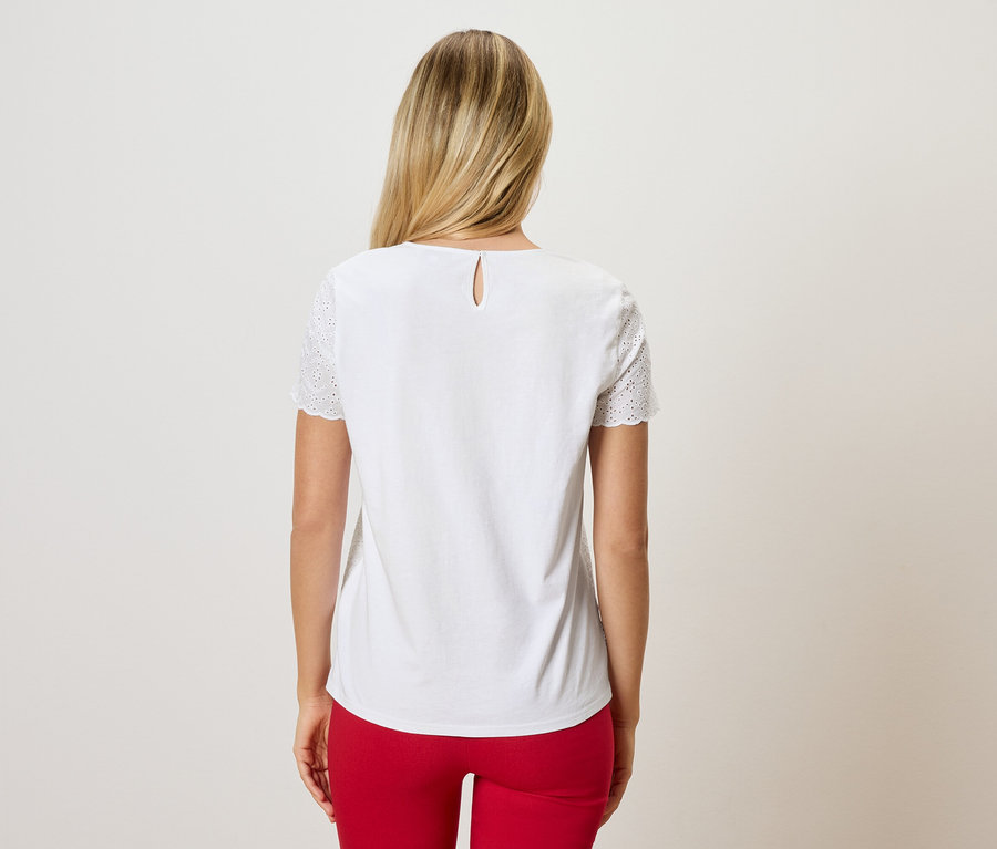 Vue de dos d'une femme aux cheveux blonds portant un chemisier blanc avec des broderies ajourées sur les manches et un pantalon rouge.