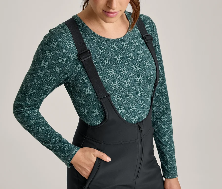 Femme portant un haut vert à manches longues avec un motif géométrique et un pantalon de ski noir à bretelles, la main dans une poche.