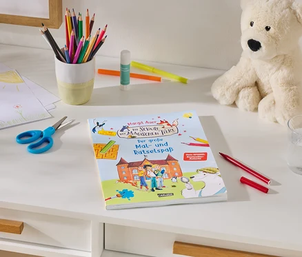 Un bureau blanc avec des fournitures d'art : une pile de dessins d'enfants, des ciseaux bleus, un pot de crayons de couleur, un bâton de colle et des marqueurs. Au centre se trouve un livre intitulé « Margit Auer Die Schule der magischen Tiere Der große Mal- und Rätselspaß ». À droite, un ours en peluche crème est assis à côté d'un verre d'eau et d'un marqueur rouge.