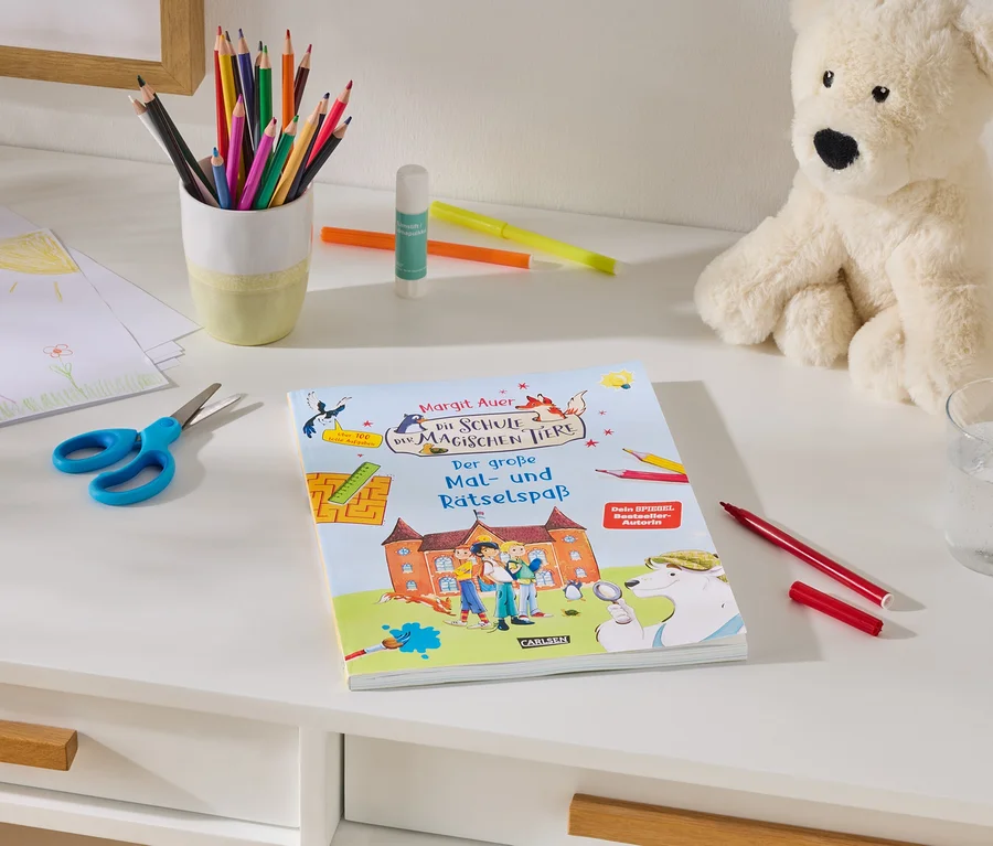 Un bureau blanc avec des fournitures d'art : une pile de dessins d'enfants, des ciseaux bleus, un pot de crayons de couleur, un bâton de colle et des marqueurs. Au centre se trouve un livre intitulé « Margit Auer Die Schule der magischen Tiere Der große Mal- und Rätselspaß ». À droite, un ours en peluche crème est assis à côté d'un verre d'eau et d'un marqueur rouge.