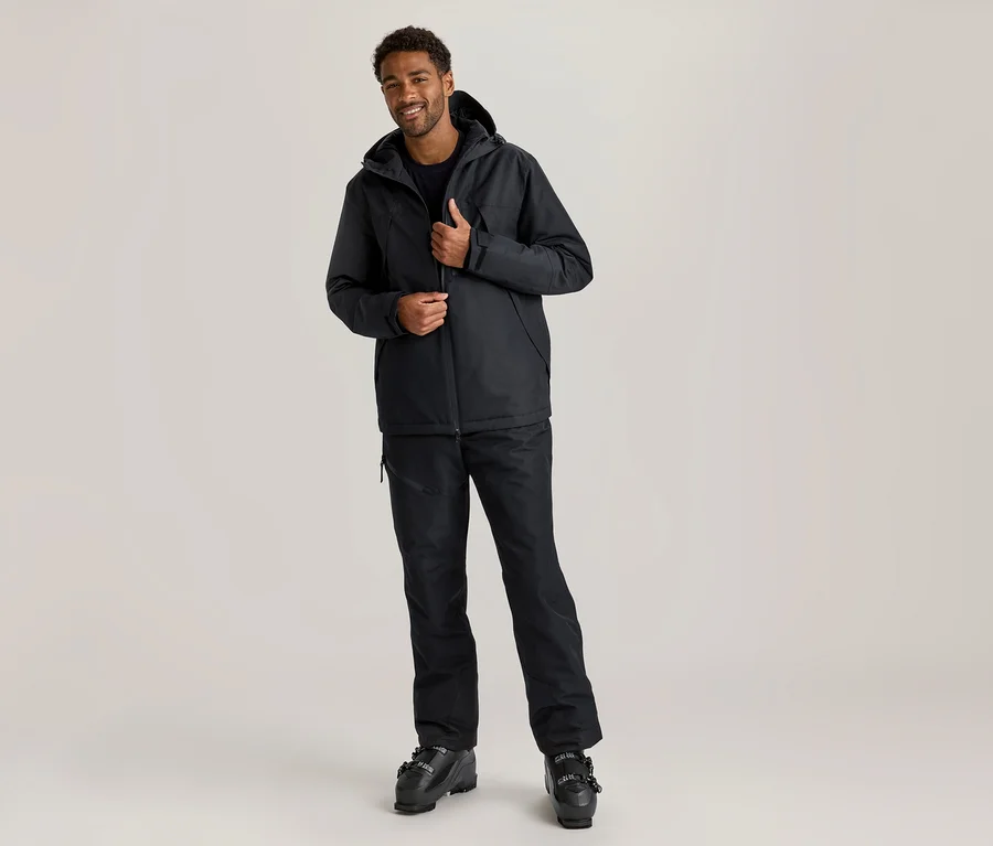 Un homme souriant à la peau foncée et aux cheveux bouclés se tient debout, ajustant sa veste de ski noire. Il porte une veste de ski noire, un pantalon de ski noir et des chaussures de ski noires.