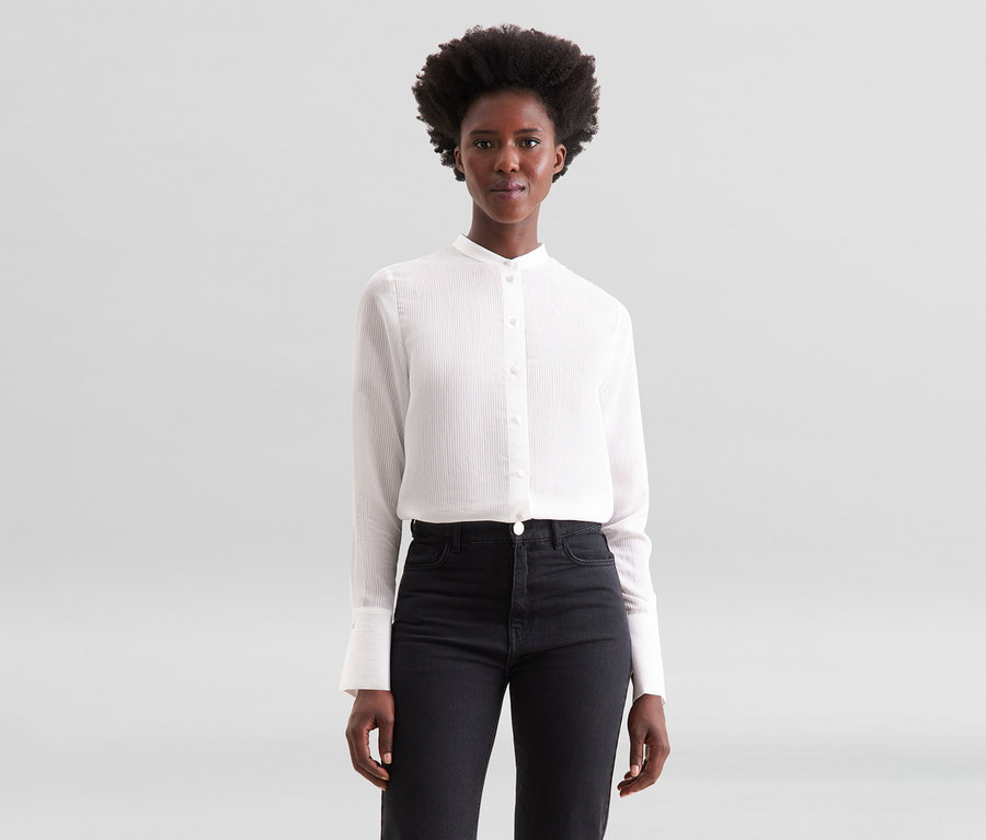 Femme aux cheveux bouclés foncés portant une chemise blanche à manches longues rayée avec un col montant et un jean noir, posant devant un fond gris clair.