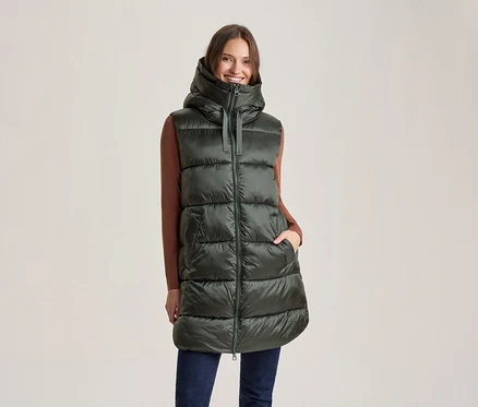 Femme souriante portant une longue doudoune sans manches vert foncé à capuche, un haut marron à manches longues et un jean bleu foncé, une main dans sa poche.