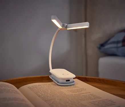 Une lampe de lecture blanche à pince éclaire un livre ouvert posé sur un plateau en bois. À l'arrière-plan, on voit un lit avec un oreiller à carreaux.