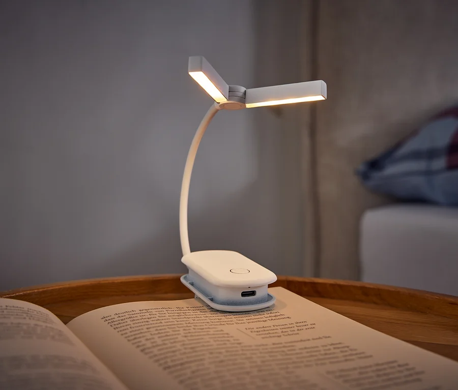 Une lampe de lecture blanche à pince éclaire un livre ouvert posé sur un plateau en bois. À l'arrière-plan, on voit un lit avec un oreiller à carreaux.
