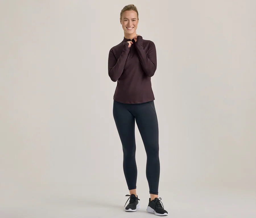Femme souriante en haut à manches longues foncé et leggings noirs, ajustant son haut avec les deux mains.