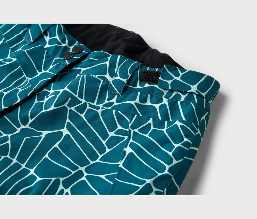 Gros plan d'un pantalon sarcelle avec un motif géométrique blanc, montrant la ceinture avec deux boutons foncés et une sangle velcro noire, ainsi qu'une doublure intérieure noire.
